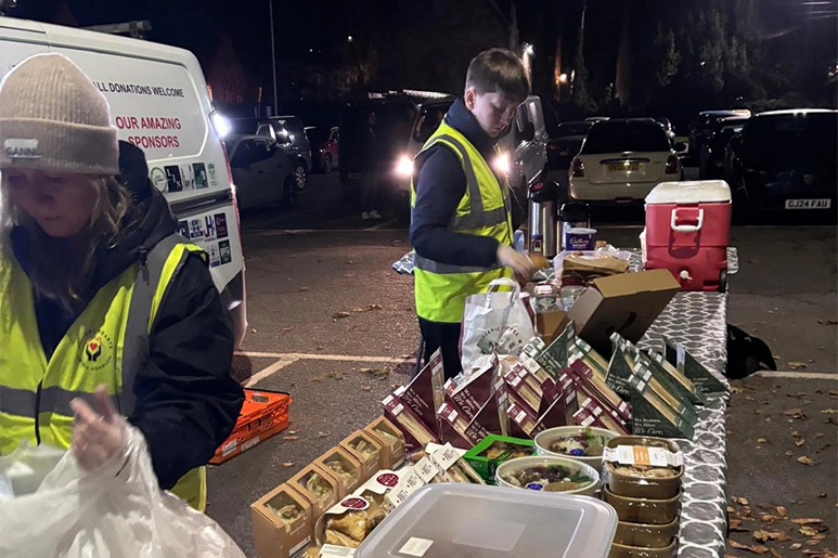 Hungry Hearts volunteers serving food at a night-time distribution table beside the donation van